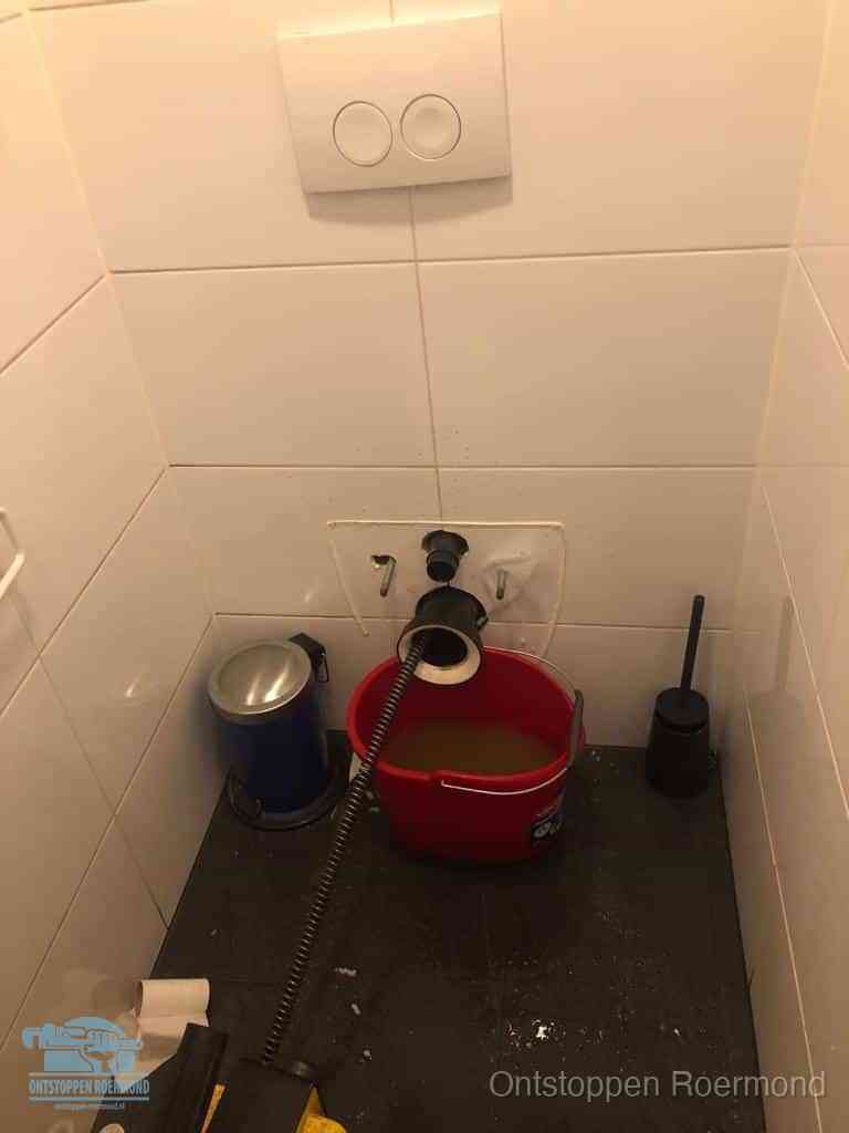 Een ontstopte wand-wc met verwijderde toiletpot waar een ontstoppingsveer in de wanduitgang verdwijnt, het achterhalen waar de verstopping zit in Roermond