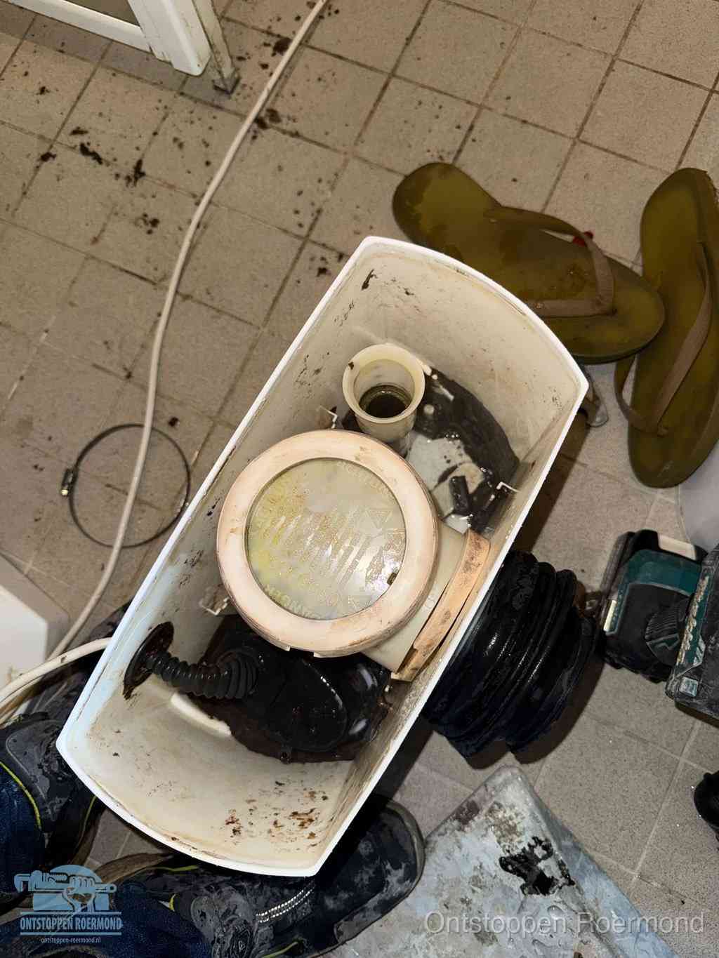 Een gedemonteerde toiletpot op de vloer met ontstoppingsveer en gereedschap ernaast, het verwijderen van een vastzittend voorwerp uit de wc in Roermond