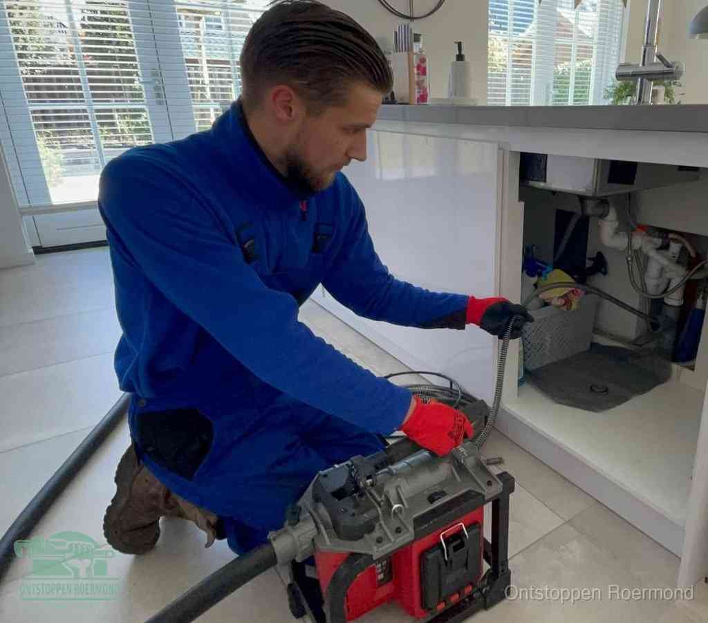 Een monteur met rode handschoenen knielt in een keuken en voert een elektrische ontstoppingsveer in tijdens het professioneel afvoer ontstoppen in Roermond